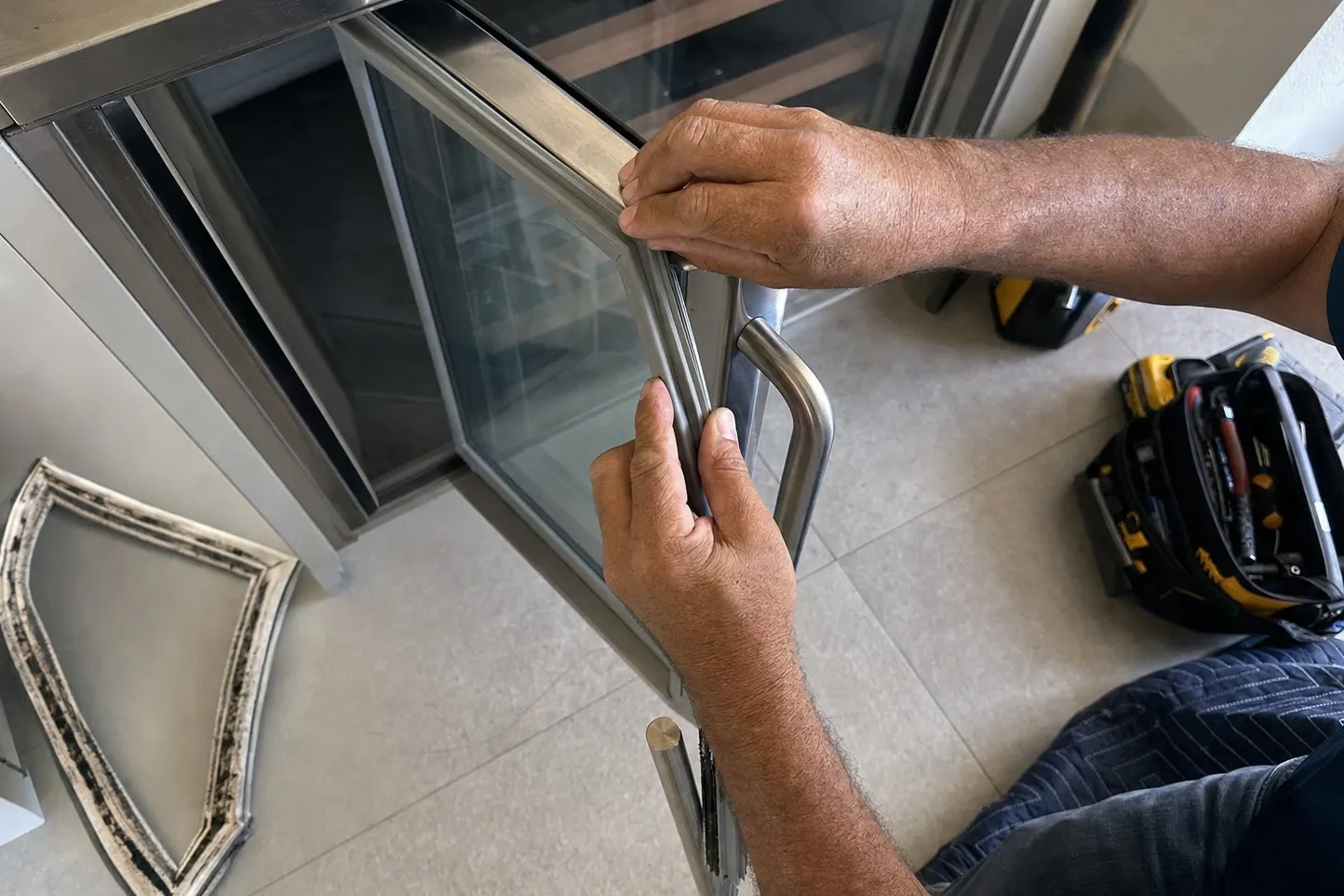 Hands easing a flexible door seal onto the glass door of an undercounter cabinet, the old seal on the floor